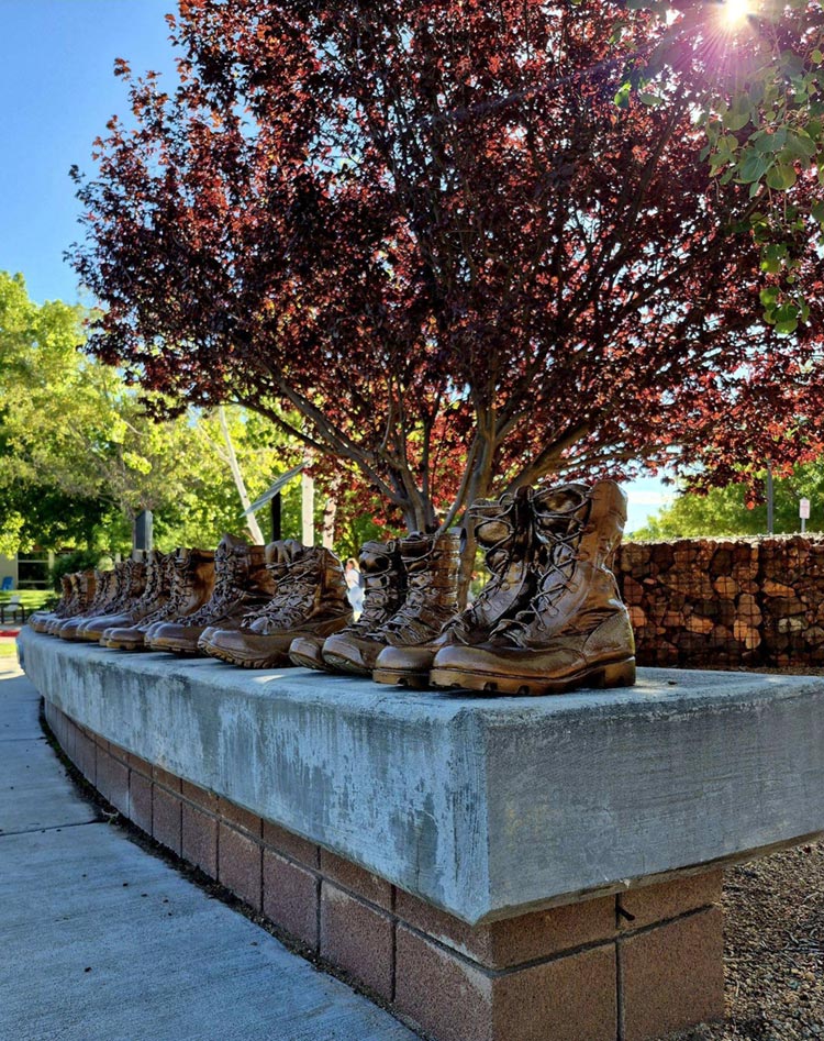 A row of memorial boots follows the curve of the stone bench beneath a tree.